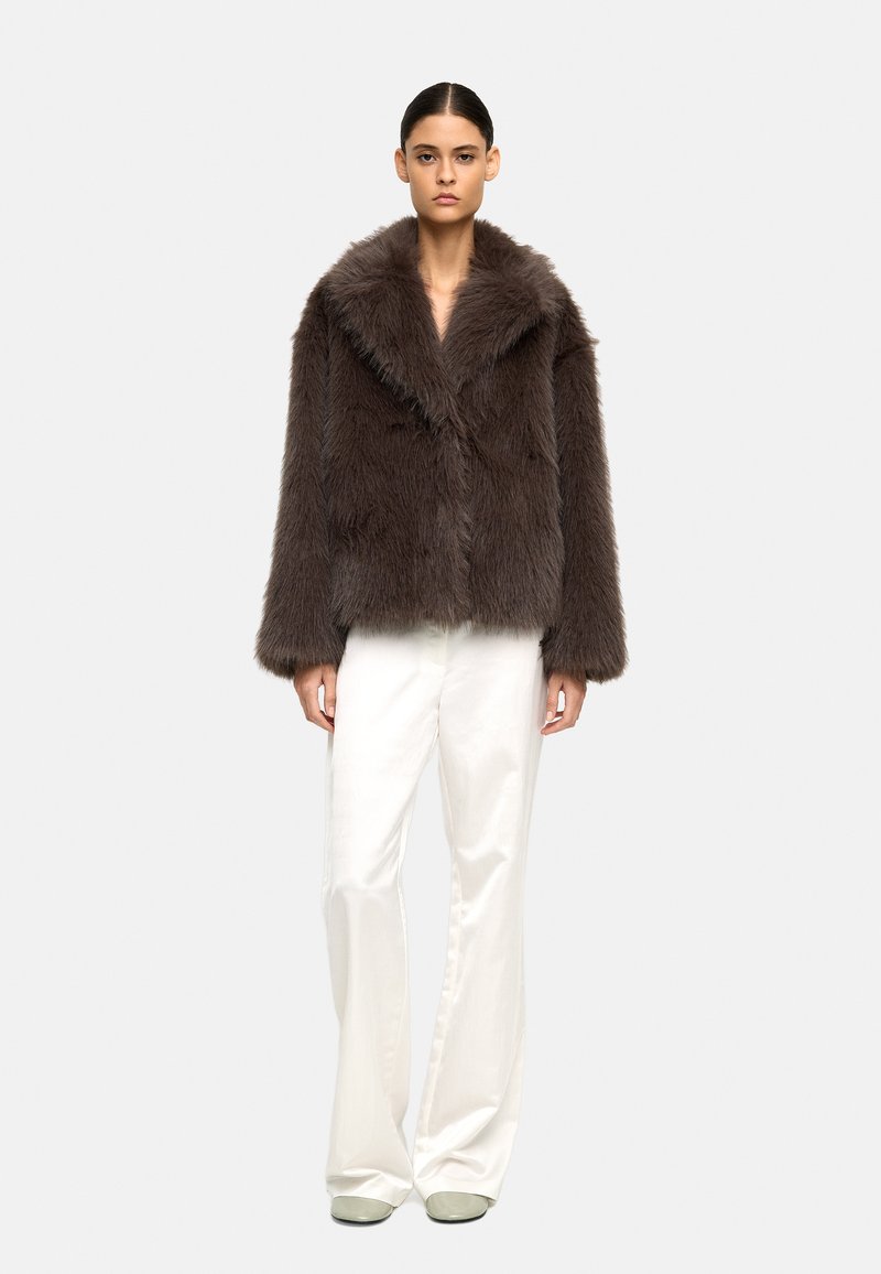Brązowa kurtka z sztucznego futra z szalowym kołnierzem, o luźnym kroju i długich rękawach, zestawiona z białymi szerokimi spodniami.