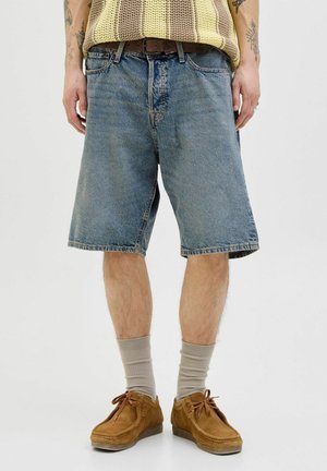 Person iført løse blå denimshorts, et brunt bælte, brune ruskindssko med beige sokker og en beige og brun stribet strikket top, stående.