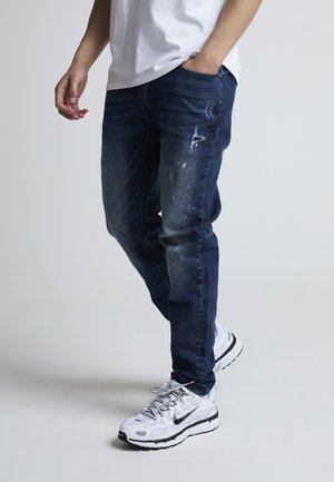 Mann iført mørkeblå, slitte jeans og hvite Nike-sneakers, stående med en hånd i lomma mot en ensfarget bakgrunn.