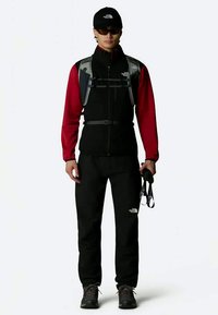 Crna vanjska prsluk s reflektirajućim detaljima preko crvene majice dugih rukava, u kombinaciji s crnim hlačama i kapom. Uključuje trakice i ruksak za hidrataciju.