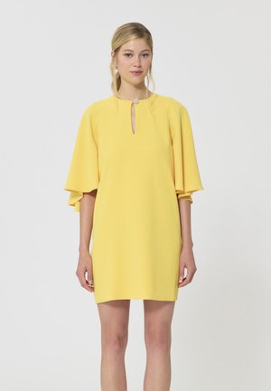 Jeune femme portant une courte robe jaune avec de larges manches évasées et un petit décolleté en forme de goutte orné d'un bouton.