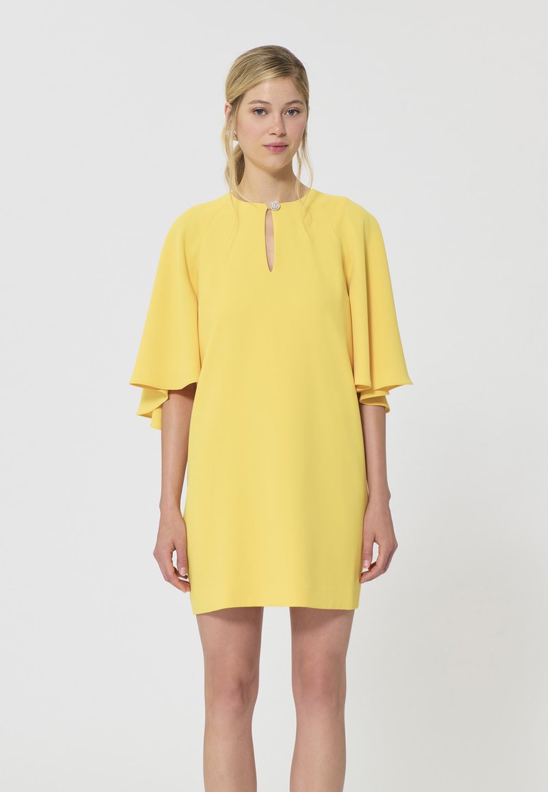 Jeune femme portant une courte robe jaune avec de larges manches évasées et un petit décolleté en forme de goutte orné d'un bouton.