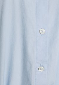 Ljusblå skjorta med knappar, som har en mjuk textur, klassisk krage och vita plastknappar fram. Subtila sömmar synliga.