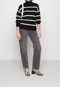 Personne portant un pull rayé noir et blanc, un jean gris, des bottines motif zèbre, et un sac à bandoulière vert clair sur un fond uni.