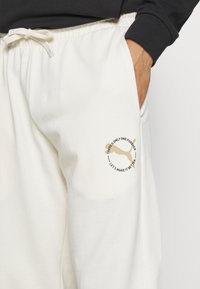 Person som bär off-white sweatpants med elastisk midja och dragsko, med en beige Puma-logotyp och texten "Det finns bara en för evigt."