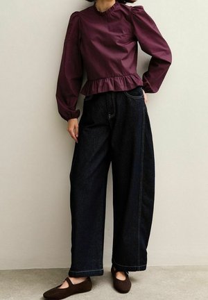 Kobieta ubrana w bluzkę z długim rękawem w ciemnym fiolecie z falbaną na dole, szerokie spodnie dżinsowe w ciemnym kolorze oraz brązowe baleriny, stojąca na tle jednolitej ściany.