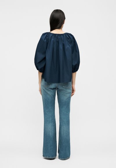 Žena s dlhými tmavými vlasmi, oblečená v námornícky modrej blúzke s nafúknutými rukávmi a svetlomodrých zvonových džínsoch, stojaca chrbtom k divákovi na jednoduchom pozadí.