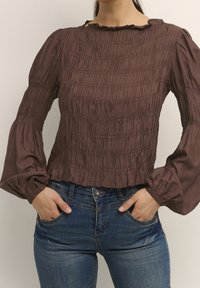 Blusa marrone a maniche lunghe con texture arricciata e scollo a volant, abbinata a jeans blu in denim, mani in tasca.
