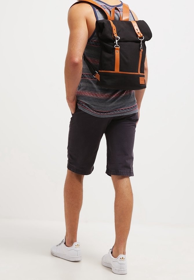 Svart canvasryggsäck med bruna läderremmar och detaljer, rektangulär form, silverfärgat hänglås, rullad topp och justerbara axelremmar.