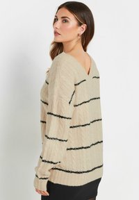 Maglione beige lavorato a maglia con strisce verticali nere, scollo a V, vestibilità comoda e dettagli a coste testurizzati. Materiale morbido e maniche lunghe.