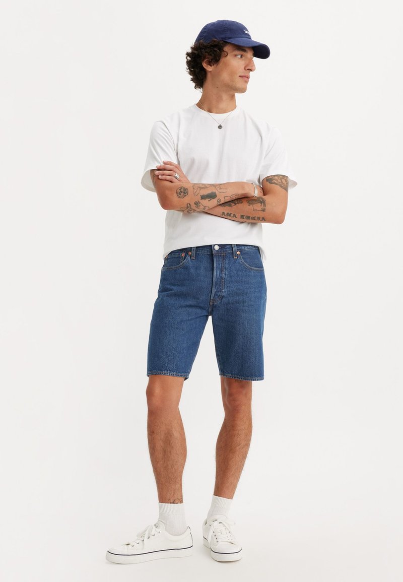 Denim shorts in blue, featuring a classic cut and raw hem. Paired with a white crew-neck t-shirt and white sneakers. Simple design.