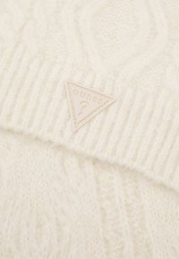 Beanie lavorato a maglia di colore crema con design a coste e motivo a treccia testurizzato. Include un patch triangolare in rilievo con il logo GUESS.