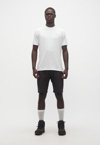 Άνδρας που φοράει λευκό μπλουζάκι, μαύρα cargo σορτς, λευκές κάλτσες πλήρους μήκους, μαύρα ορειβατικά μποτάκια και σκούρα γυαλιά ηλίου, στέκεται μπροστά σε λευκό απλό φόντο.