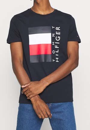 Osoba w ciemnej koszulce z dużym logo Tommy Hilfiger i dżinsach stoi z założonymi rękoma na gładkim, jasnym tle.