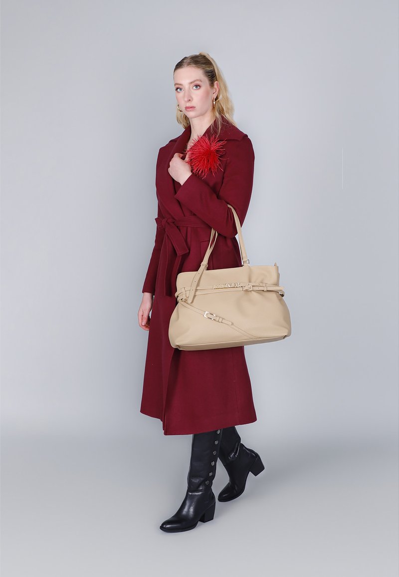 Beige handtas met gestructureerd materiaal en verstelbare riemen, gedragen met een bordeauxrode jas; model in zwarte kniehoge laarzen met knopen.