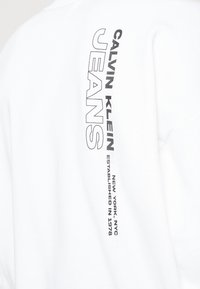 Sudadera blanca con un diseño de texto vertical que presenta "CALVIN KLEIN JEANS" en letras negras en negrita, detallando "NUEVA YORK NYC ESTABLECIDO EN 1978."