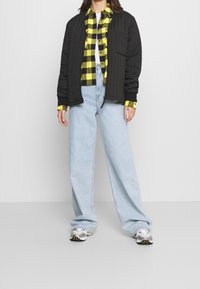 Person wearing black quilted jacket over yellow and black plaid shirt, light blue wide-leg jeans, and white sneakers standing on gray floor.