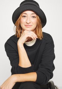 Jeune femme aux cheveux roux courts portant un chapeau noir et un pull, reposant son menton sur sa main, assise dans un fauteuil roulant sur un fond blanc.