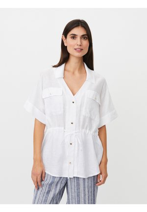 Mujer que lleva una camisa blanca de manga corta con botones, bolsillos en el pecho y cinturón a la cintura, combinada con pantalones a rayas azules y blancas.