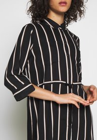 Abito camicia a righe bianche e nere con colletto, maniche corte, bottoni frontali e dettaglio in vita legato. Il tessuto sembra leggero e liscio.