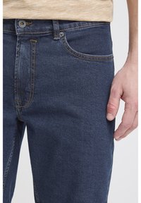 Tummansiniset farkut, joissa näkyvät ompeleet, etutasku ja nappikiinnitys. Sileä pinta ja kapeutettu malli.