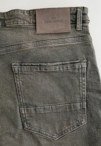 Poche arrière d’un jean en denim noir délavé avec des détails cousus et un patch en cuir portant l’inscription « NEXT DENIM SUPPLY » au-dessus de la ceinture.