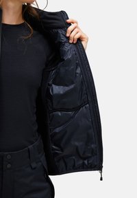 Zwarte buitenjas met een gladde, glanzende voering. Voorzien van een ritssluiting en zichtbare zakken met een strakke, minimalistische uitstraling.