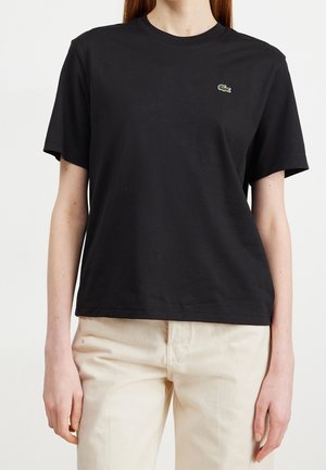 Une personne portant un t-shirt noir uni à manches courtes avec un petit logo de crocodile vert sur la poitrine gauche, associé à un pantalon beige clair.