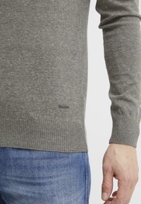 Pulover gri tricotat cu manșete și tiv cu dungi, având o etichetă mică și închisă la culoare pe partea inferioară. Purtați-l peste blugi din denim albastri.