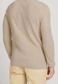 Maglione beige a coste con lavorazione a maglia strutturata. Presenta maniche lunghe e orlo arrotondato. Indossato sopra pantaloni chiari.