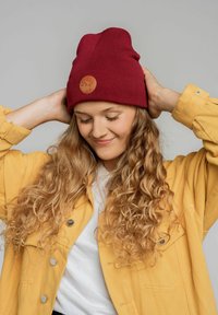 Bordowy, dziany beanie o teksturowanym wykończeniu z okrągłą naszywką z logo. Uzupełnione żółtą kurtką i białą koszulą.