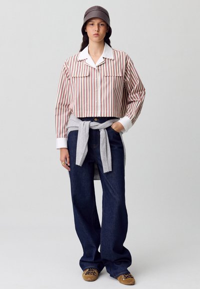 Camisa de rayas corta con dos bolsillos frontales, combinada con pantalones de mezclilla oscuros y holgados, y un suéter gris atado a la cintura; zapatos marrones con cordones.