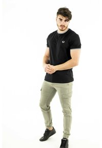 Sort kortærmet t-shirt med hvid kant og logo, parret med lysegrønne cargo-bukser og sorte sneakers, mod en hvid baggrund.