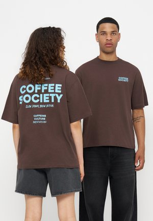Une femme et un homme portent des t-shirts marron « Coffee Society » avec un texte bleu ; la femme est de dos, l’homme regarde devant avec une expression neutre.
