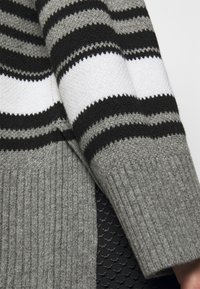 Szary sweter dzianinowy w czarno-białe paski, z ściągaczami na rękawach i teksturowanym wykończeniem. Akcenty obejmują różne szerokości pasków.