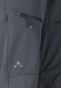 Mørkegråt stof med en lynlåslomme med en sort lynlåstrækker og et lille broderet trekantet logo nær sømmen.