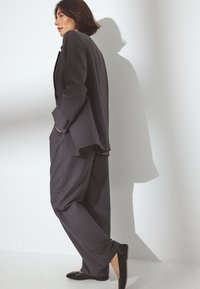 Priešingas tamsiai pilkas kostiumas, turintis laisvai priglūstančią švarką ir plačius kelnes. Papildytas juodais butais. Lygus audinio paviršius ir klasikinis dizainas.