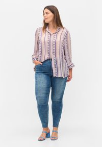 Blusa ligera y estampada con rayas moradas y rosas, combinada con jeans azules. La modelo lleva sandalias de tacón azules y tiene el cabello largo y castaño.