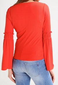 Röd ribbad långärmad topp med utsvängda ärmar och rynkade detaljer vid handleden, kombinerad med ljusblå denimjeans.