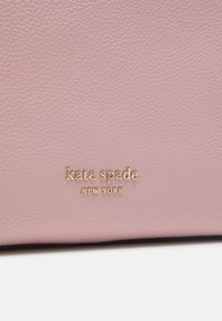 Rosa texturerad läder med en guldfolie "kate spade NEW YORK"-logotyp. Ytan visar ett prickigt mönster som tillför visuell intresse.