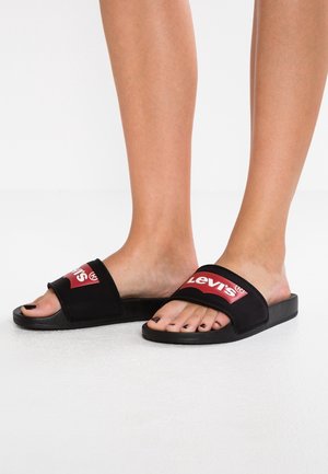 Jalat, joissa on mustat Levi's-liukusandaalit, joissa on punavalkoinen logo hihnoissa, seisovat sileällä valkoisella pinnalla.