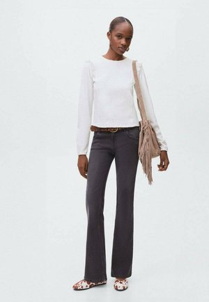 Kvinde iført en hvid langærmet top, mørke flagrende jeans, brun bælte, mønstrede sko og bærende en beige skuldertaske med frynser.
