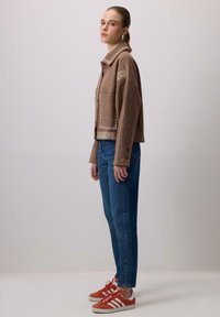 Hnedá skrátená bunda s vyšívanými detailmi, skombinovaná s džínsami s vysokým pásom a červenými teniskami s bielymi pruhmi.