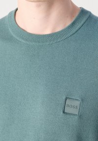 Gros plan d'une personne portant un pull col rond bleu sarcelle avec un petit écusson carré avec le logo « BOSS » sur la poitrine.