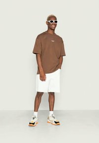 T-shirt castanho de mangas curtas com logótipo branco, combinado com calções brancos. O modelo usa óculos de sol brancos e sapatilhas robustas com detalhes em preto e laranja.