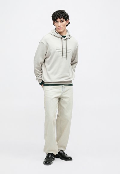 Φωτεινό γκρι φούτερ με ανάγλυφη λεπτομέρεια λογότυπου, σε συνδυασμό με μπεζ τζιν φαρδιάς γραμμής και μαύρα παπούτσια. Μινιμαλιστικός σχεδιασμός και μαλακές υφές υφάσματος.