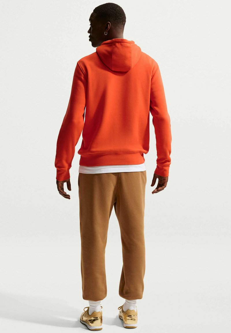 Sudadera con capucha naranja, puños y dobladillo acanalados. Combinada con pantalones marrones estrechos y zapatillas doradas. Vista trasera que destaca un diseño sencillo.