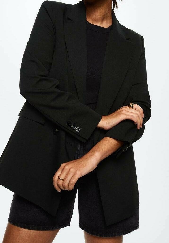 Personne portant un blazer noir oversize sur un top noir et un short noir, avec des bagues en or aux doigts, bras croisés.