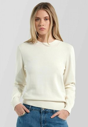 Giovane donna con capelli biondi lisci indossa un maglione bianco a maniche lunghe semplice e jeans blu, mani nelle tasche, sfondo neutro.