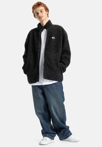 Zwarte fleecjas met hoge kraag en ritssluiting, voorzien van twee grote voorkruiskosten en een logo patch. Draagt over een wit shirt en jeans.